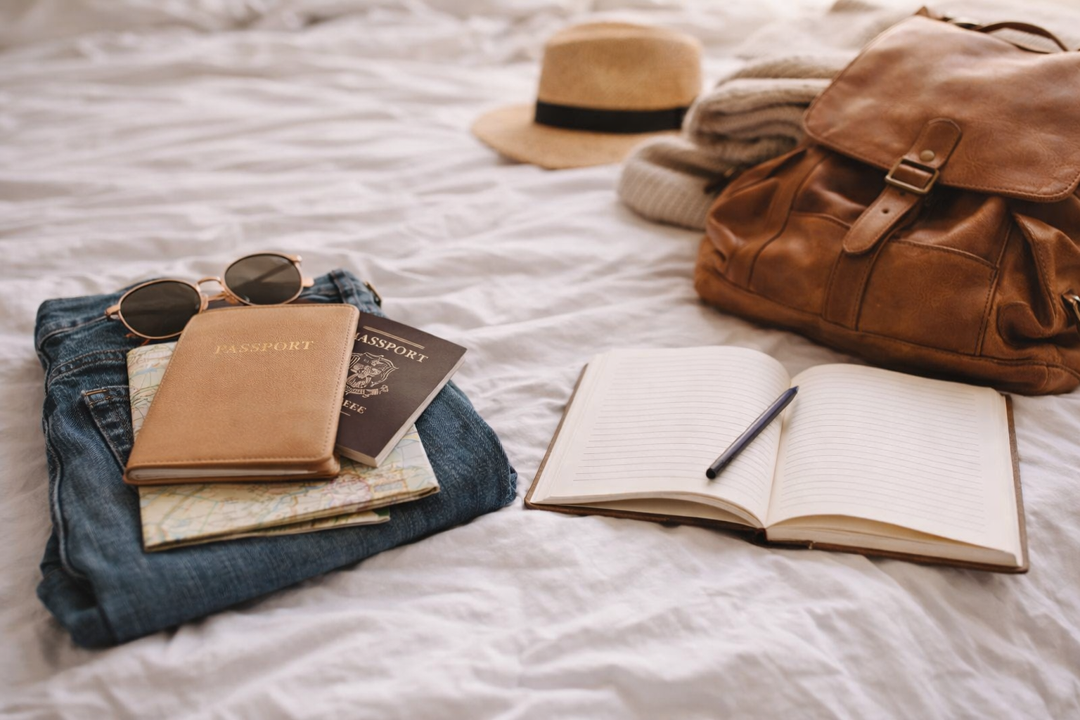 Travel essentials laid out on a bed in soft natural light, including a leather backpack, passport, notebook, sunglasses, map, and folded clothes, representing intentional travel and preparation.