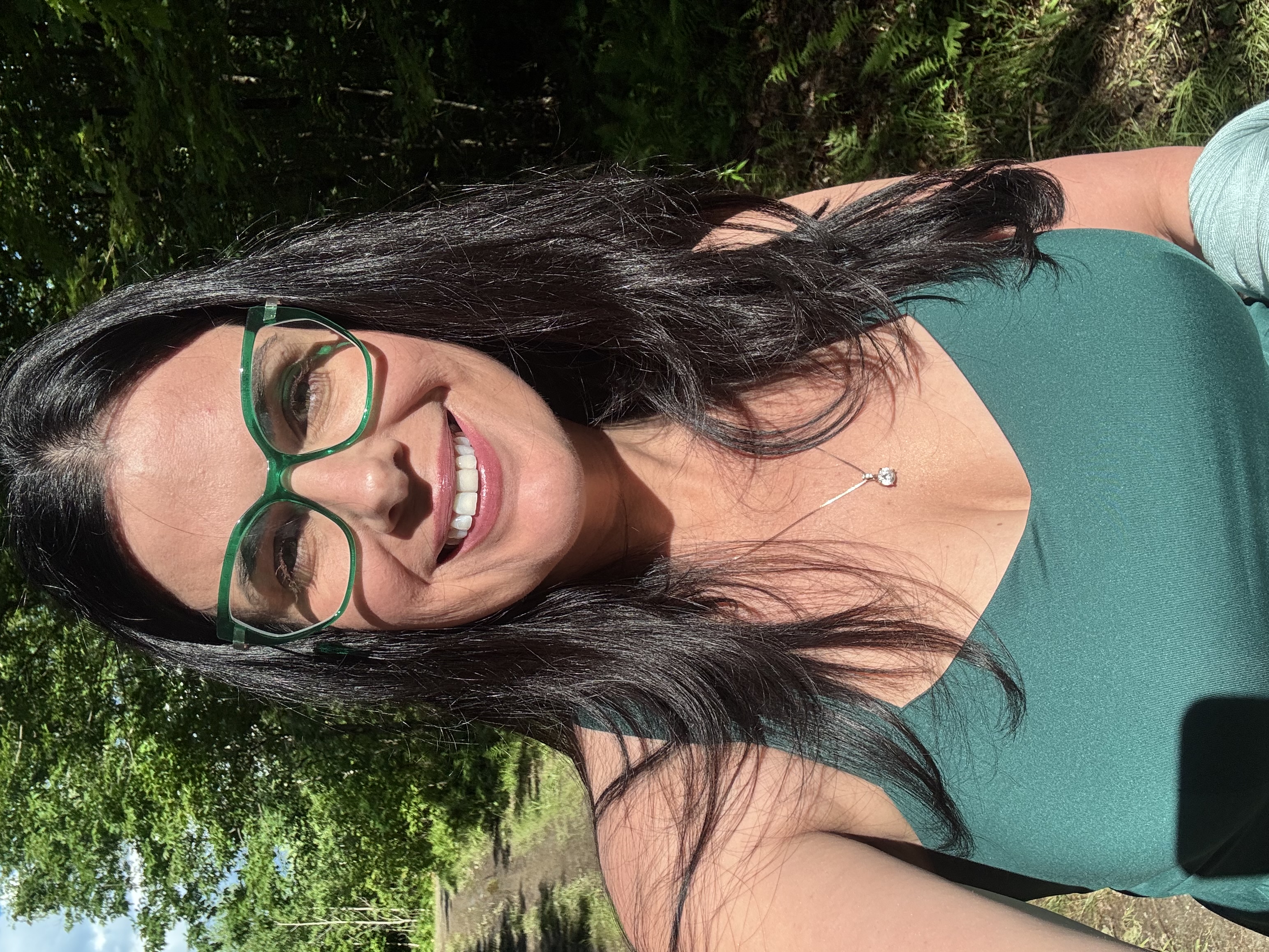 A smiling woman standing on a wooded trail in the morning sunlight, reflecting on how small daily actions build a meaningful life over time.